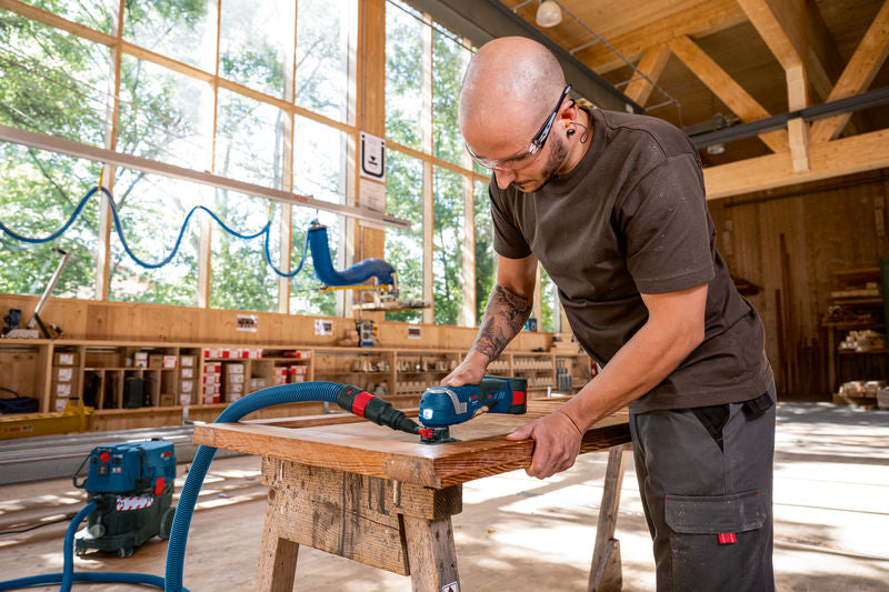 Eine Person mit Schutzausrüstung schleift mit einem Elektrowerkzeug Holz auf einer Werkbank.