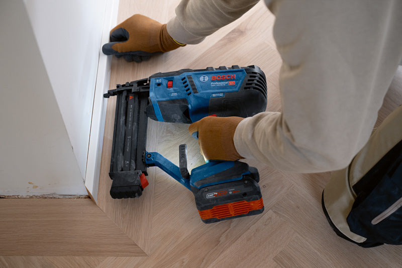 Eine Person mit Sicherheitsausrüstung befestigt mit einem Akku-Holznagler Zierleisten am Boden.