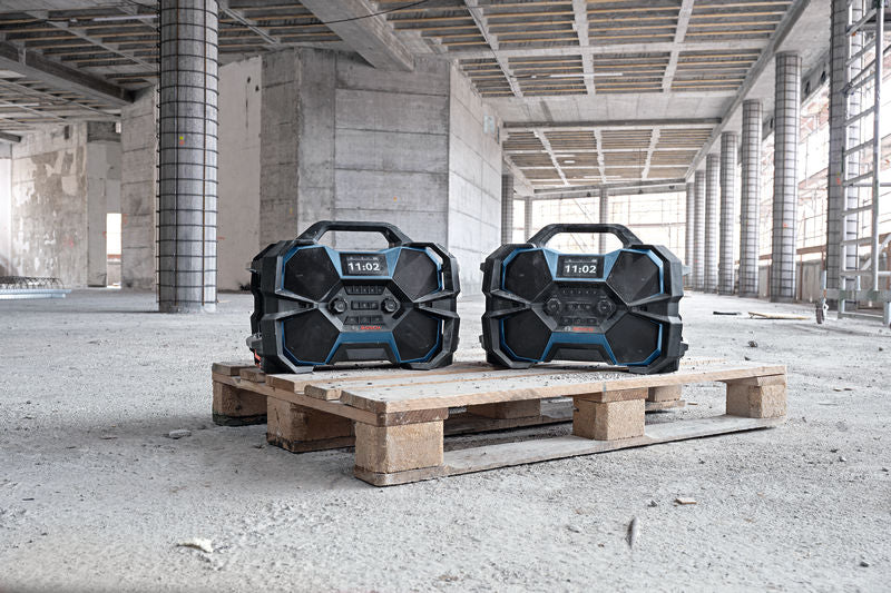 Zwei robuste Baustellenradios auf einer Holzpalette in einer teilweise errichteten Struktur.
