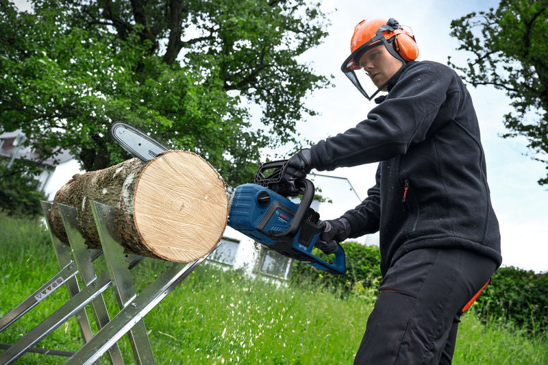 Eine Person mit Sicherheitsausrüstung verwendet eine Akku-Kettensäge, um im Freien einen Baumstamm zu sägen.