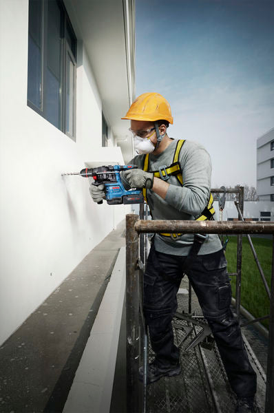 Eine Person mit Sicherheitsausrüstung bohrt von einem Gerüst aus in eine Gebäudewand.