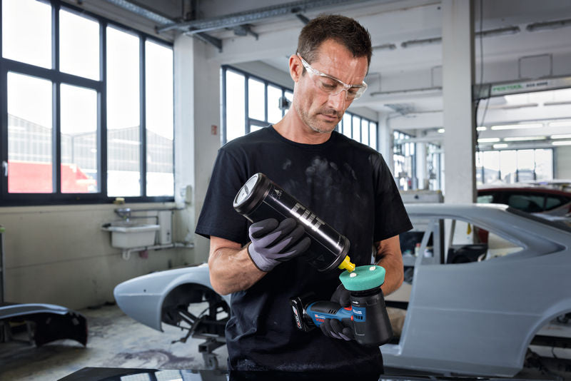 Eine Person mit Sicherheitsausrüstung trägt in einer Autowerkstatt Politur auf einer Poliermaschine auf.