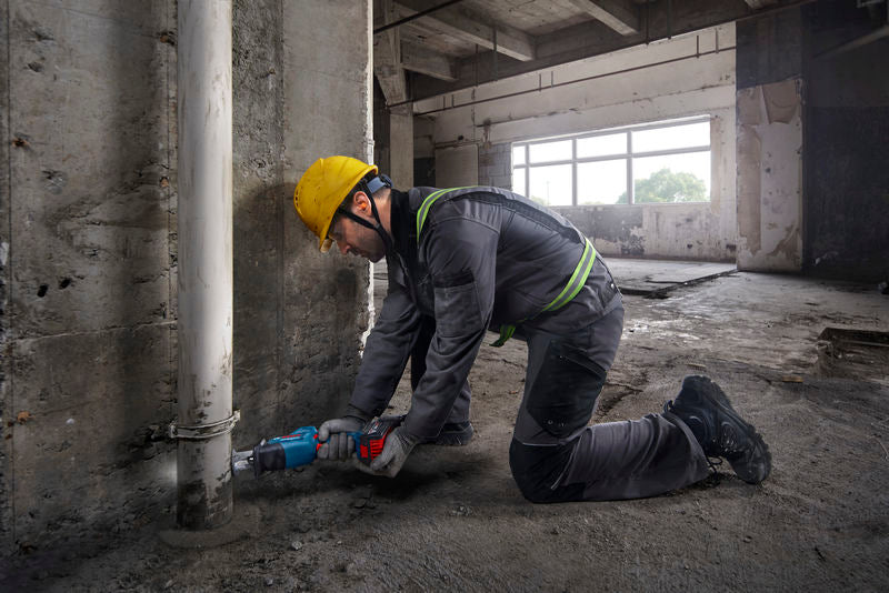 Eine Person mit Sicherheitsausrüstung schneidet mit einer Säbelsäge ein Rohr in der Nähe einer Betonwand durch.