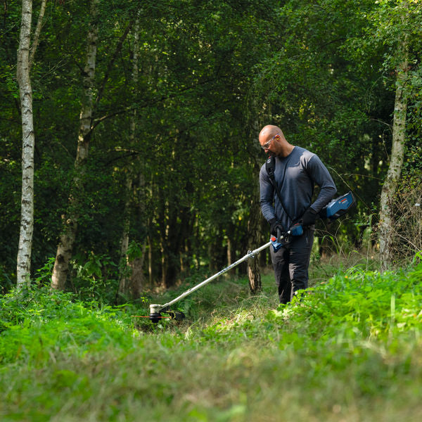 Eine Person mit Sicherheitsausrüstung schneidet in einem Wald Gras mit einem Akku-Rasentrimmer.