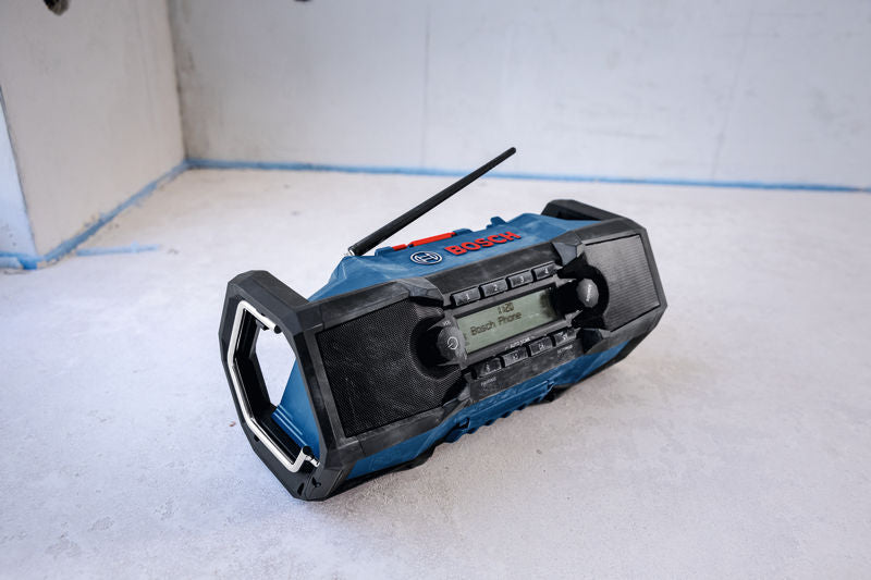 Ein tragbares Baustellenradio mit Display und zwei Lautsprechern steht auf einem Betonboden.
