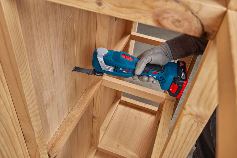 Eine Person mit Schutzausrüstung schneidet mit einem Multifunktionswerkzeug eine rechteckige Öffnung in einen Holzschrank.