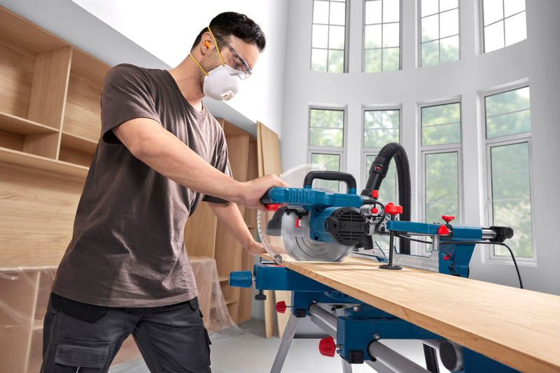 Eine Person in Schutzausrüstung schneidet Holz mit einer Gehrungssäge in einer hellen Werkstatt.