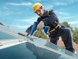 Eine Person mit Sicherheitsausrüstung zieht mit einem Elektrowerkzeug eine Schraube an einem Solarpanel auf dem Dach fest.
