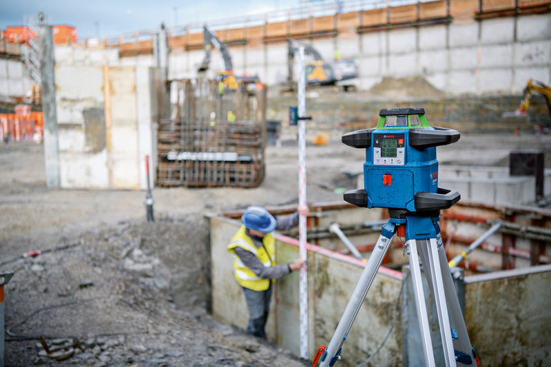Lasernivelliergerät auf einem Stativ auf einer Baustelle, Person trägt Sicherheitsausrüstung.
