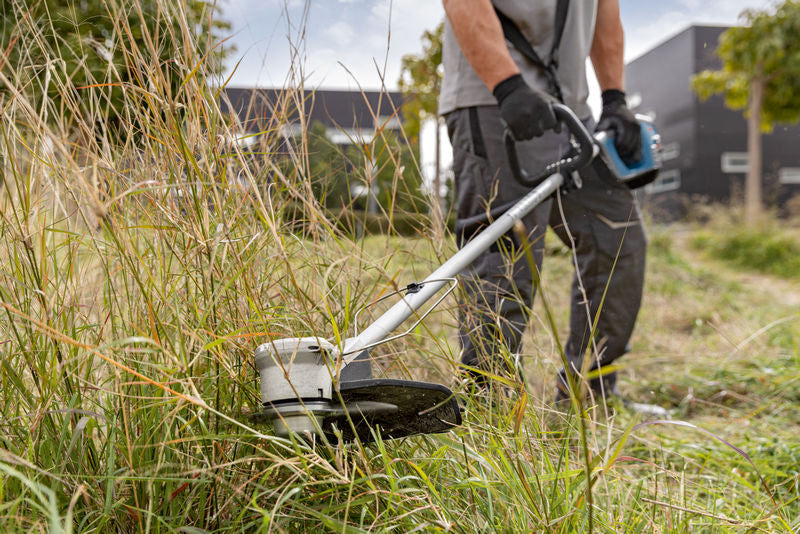 Eine Person mit Schutzausrüstung schneidet hohes Gras mit einer Akku-Motorsense.