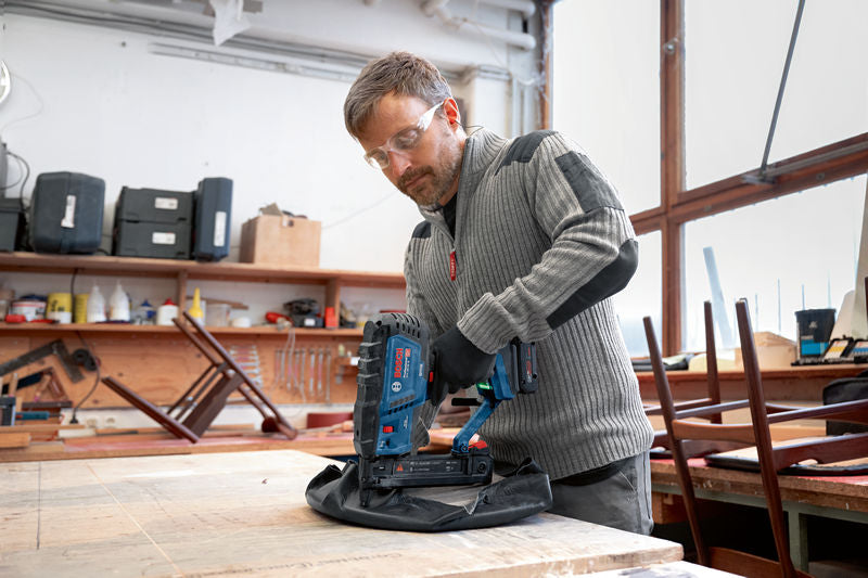 Eine Person mit Sicherheitsausrüstung heftet mit einem Akku-Holztacker Stoff auf Holz.