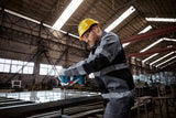 Eine Person mit Sicherheitsausrüstung verwendet eine Bohrmaschine an einem Metallrahmen in einer Fabrik.