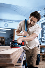 Eine Person mit Schutzausrüstung schleift Holzbretter mit einem Akku-Exzenterschleifer