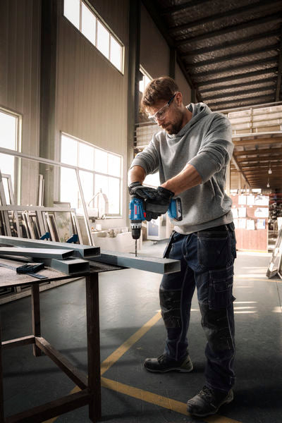 Eine Person mit Schutzausrüstung bohrt in einer Werkstatt in einen Metallträger.
