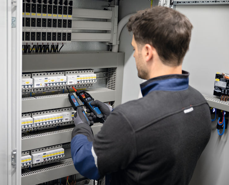 Eine Person mit Sicherheitsausrüstung verwendet einen Spannungsprüfer an einer Schalttafel