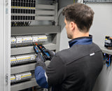 Eine Person mit Sicherheitsausrüstung verwendet einen Spannungsprüfer an einer Schalttafel