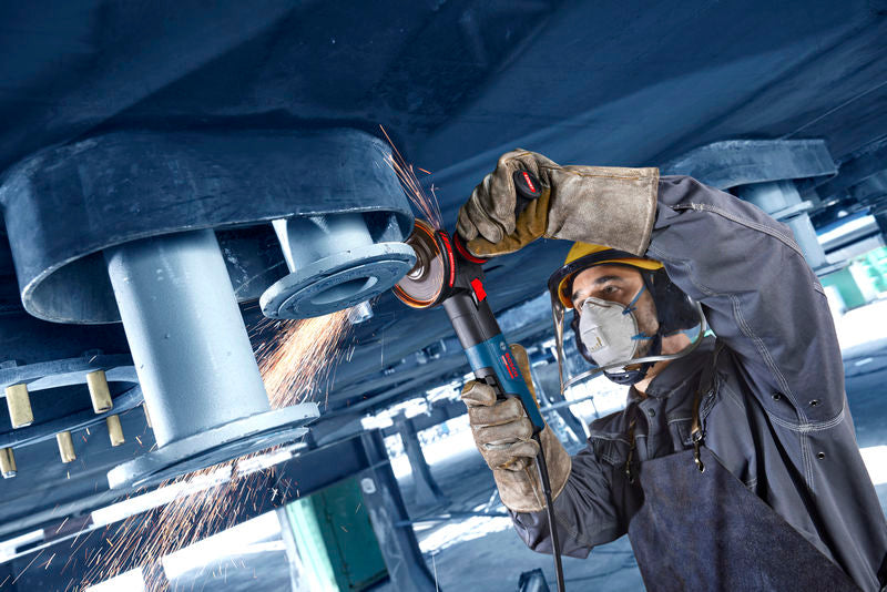 Eine Person mit Schutzausrüstung schleift mit einem Winkelschleifer über dem Kopf Metall, wodurch Funken entstehen