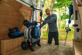 Eine Person lädt einen blauen Rasenmäher in einen Lieferwagen, der neben einem Waldstück geparkt ist.