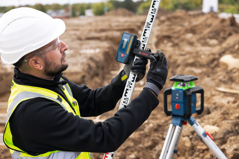 Eine Person, die Schutzausrüstung trägt, richtet auf einer Baustelle mithilfe eines Laser-Nivelliergeräts eine Nivellierlatte aus.